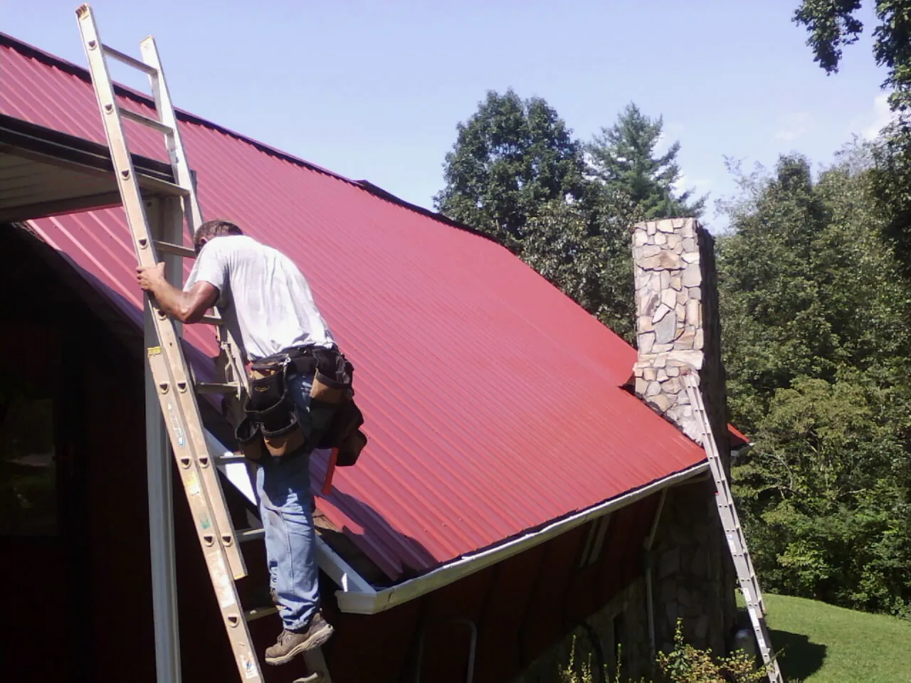 Roofer on a ladder beside a red metal roof and stone chimney — Soffit & Fascia Repair by Roofing Brothers in Hutchinson